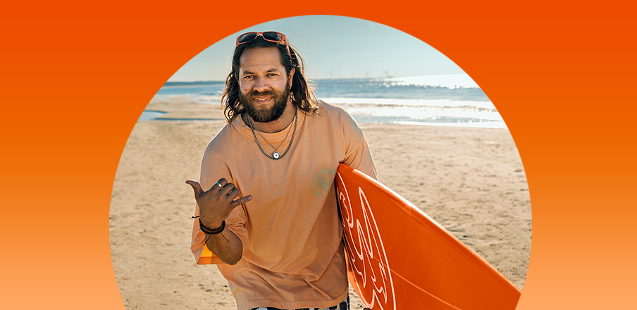 Smilende mand med langt hår og skæg på strand med orange surfbræt, viser "hang loose" håndtegn i solskinsvejr.