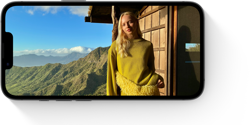 Person i gul sweater står ved træhytte med udsigt over grønne bjerge og blå himmel vist på mobilskærm.