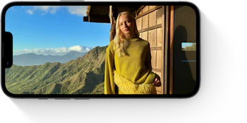 Person i gul sweater står ved træhytte med udsigt over grønne bjerge og blå himmel vist på mobilskærm.