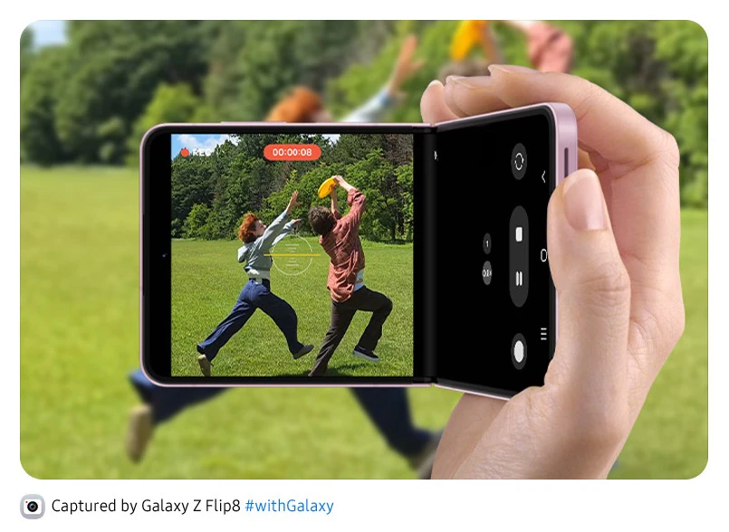 En hånd holder en pink foldbar smartphone og optager to personer, der leger med en frisbee i en grøn park.