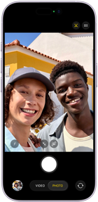 To personer tager en selfie sammen udendørs foran en gul bygning på en solrig dag.