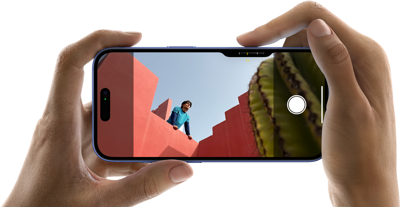 To hænder der holder en iPhone 16 vandret i hver deres ende, mobilens skærm er inde på kameraret, hvor man kan se en mand læne sig over solbrændte murer, hvor manden kigger ud over murene foran ham, i højre hjørne kan man se en kaktus stikke frem 