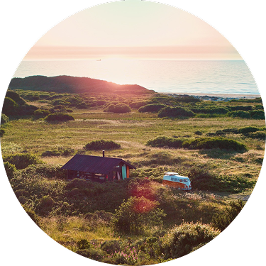 Kystlandskab ved solnedgang med en lille hytte og en klassisk VW-bus omgivet af grønne bakker og havet i baggrunden.