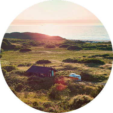 Kystlandskab ved solnedgang med en lille hytte og en klassisk VW-bus omgivet af grønne bakker og havet i baggrunden.