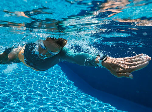Person iført svømmebriller og sportsbeklædning svømmer under vandet i en pool med smartwatch på håndleddet.