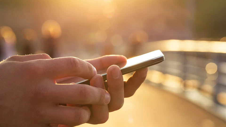 Holder en mobil i hånden med sol i baggrunden