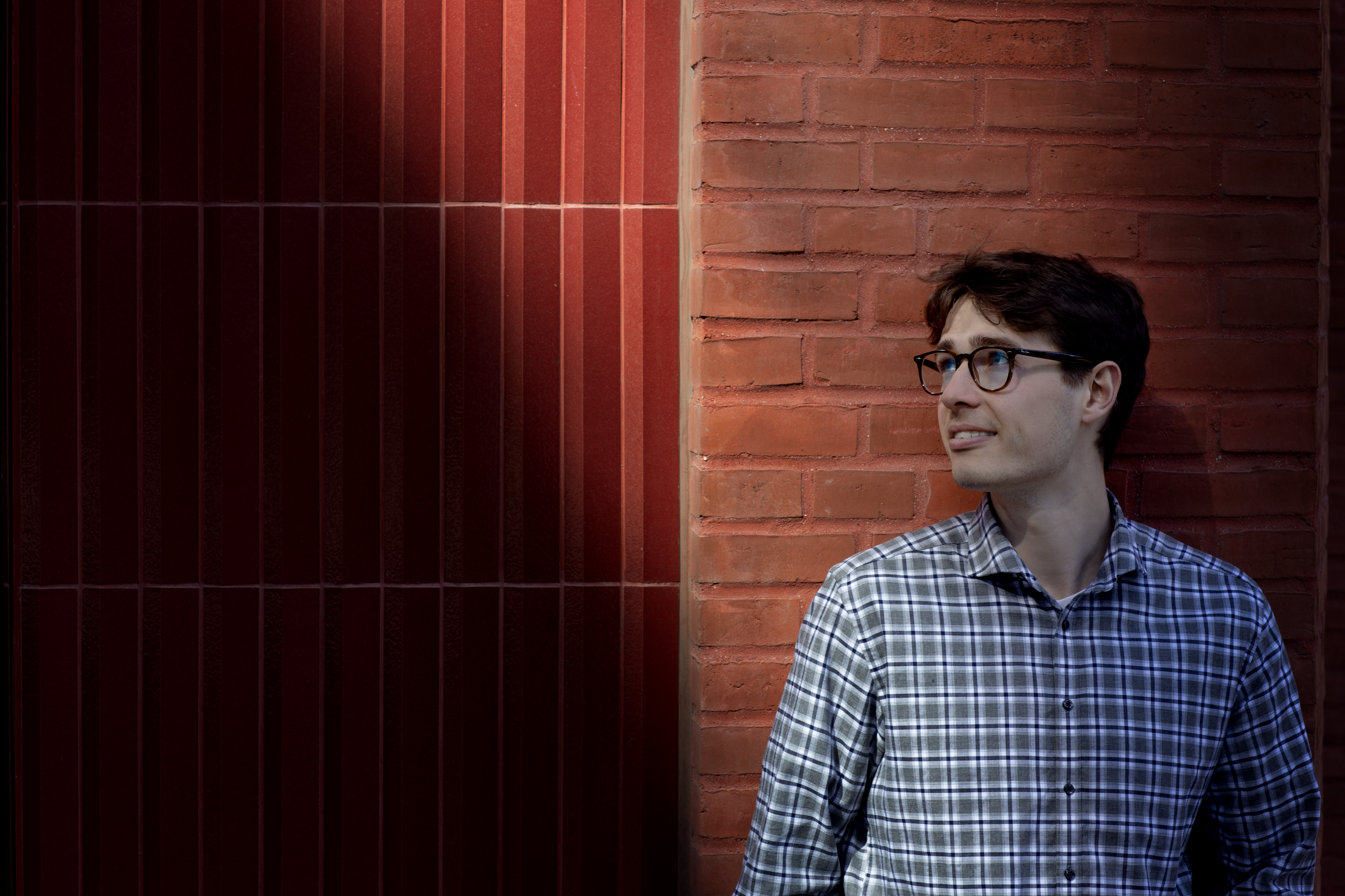 Om 3. Lukas Hvelplund - Studentermedhjælper i Customer Experience fortæller om sit arbejde i 3. Står op ad rød mur og ser til siden. 