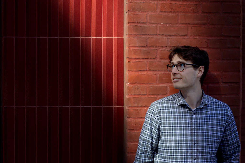 Om 3. Lukas Hvelplund - Studentermedhjælper i Customer Experience fortæller om sit arbejde i 3. Står op ad rød mur og ser til siden.